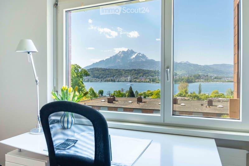 Bright room with large windows offering scenic mountain and lake views, furnished with a desk, chair, lamp, and flower vase.