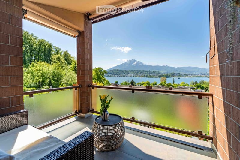 Balcony with a scenic view of a lake and mountains, featuring a small round table, a wicker chair, and a vase with yellow flowers.