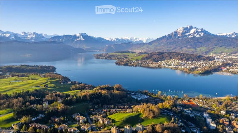 Aerial view of a scenic lakeside town surrounded by mountains, with a lake, green fields, and scattered buildings.
