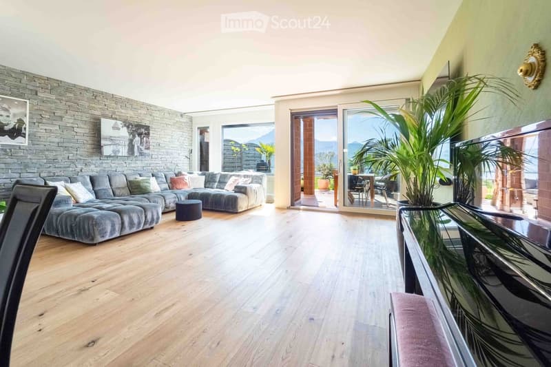 Modern living room with wooden flooring, grey sectional sofa, ottoman, glass coffee table, and large windows offering a scenic view.