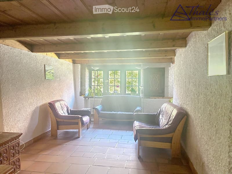 Living room with exposed wooden beams, tiled floor, and large windows. Features a sofa, two armchairs, and a small table.