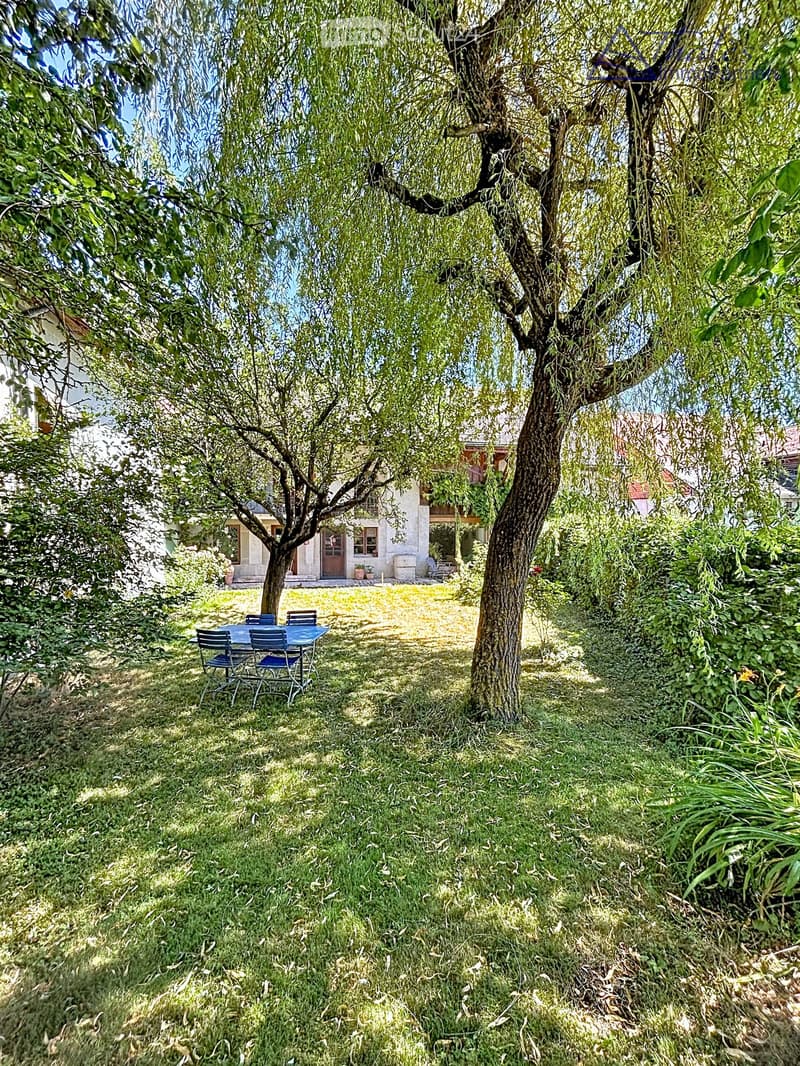 Lush garden with a large tree, a small table and chairs, and a house in the background.