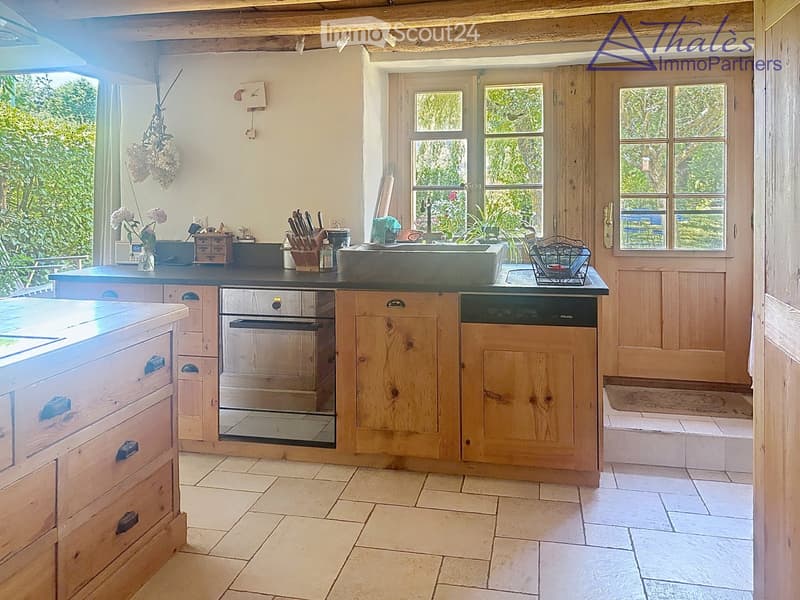 Bright kitchen with wooden cabinets, stainless steel appliances, and a large sink. Features a tiled floor, natural light from windows, and decorative elements like a flower vase and hanging herbs.