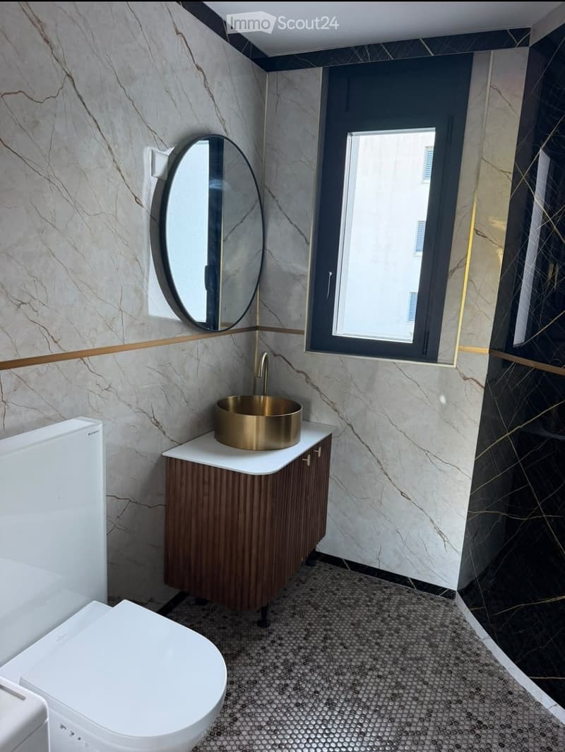 Modern bathroom with marble walls, a white toilet, a gold sink, a round mirror, and a wooden cabinet.