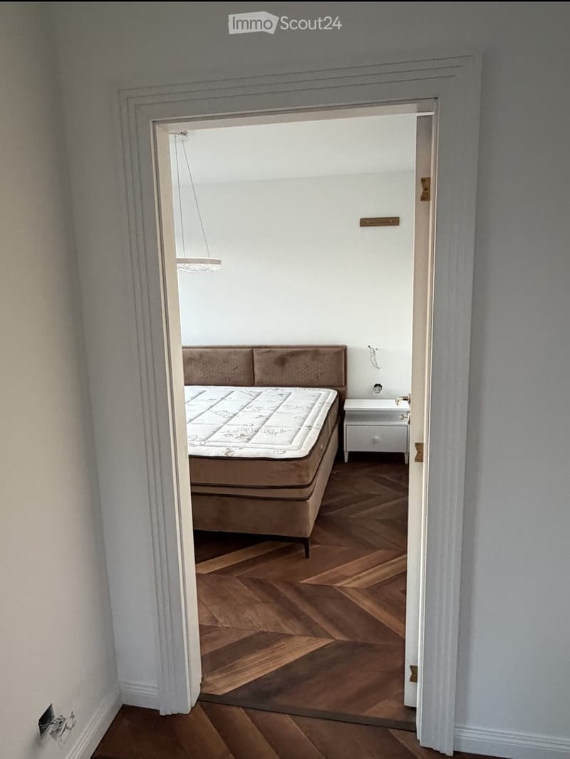 A furnished bedroom with a wooden bed, mattress, and a small nightstand. The room features wooden flooring and white walls.