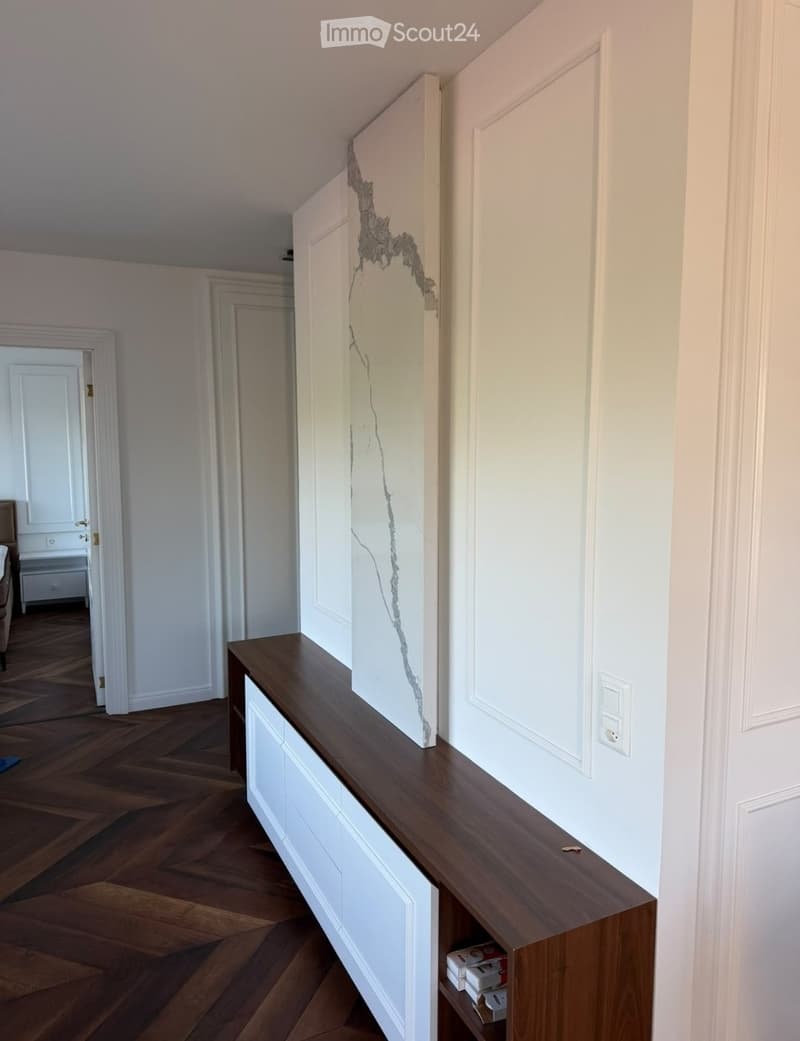 A narrow hallway with wooden flooring, white walls, and a long wooden bench with white drawers. The hallway features several doors and a visible electrical outlet on the wall.