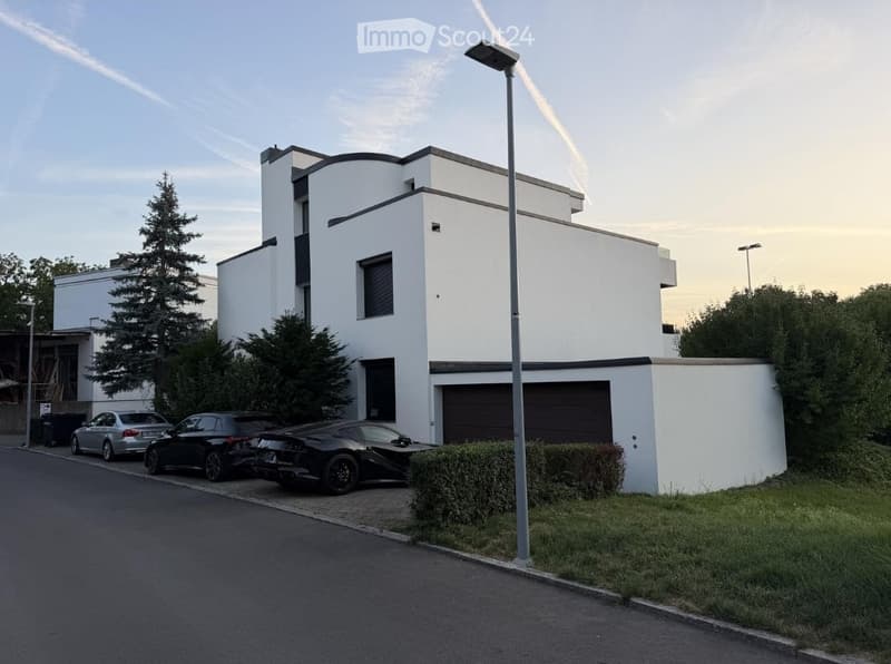 Modern two-story white building with flat roof and garage, surrounded by greenery and parked cars.