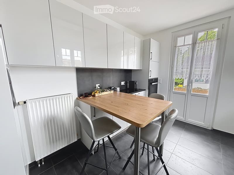Modern kitchen with white cabinets, a wooden dining table, four grey chairs, and a built-in oven and fridge.