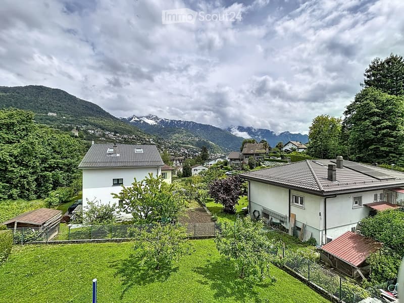 A two-story house with a tiled roof, surrounded by greenery and trees, with a scenic mountain view in the background.