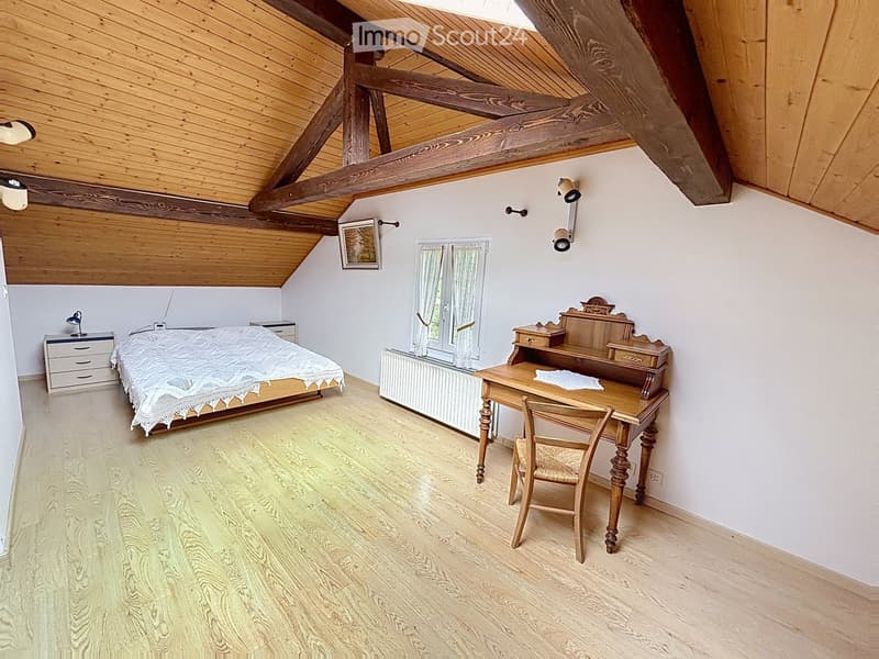Cozy attic bedroom with wooden slanted ceilings, a double bed with white bedding, a wooden desk with a chair, a nightstand, and a window with white curtains.