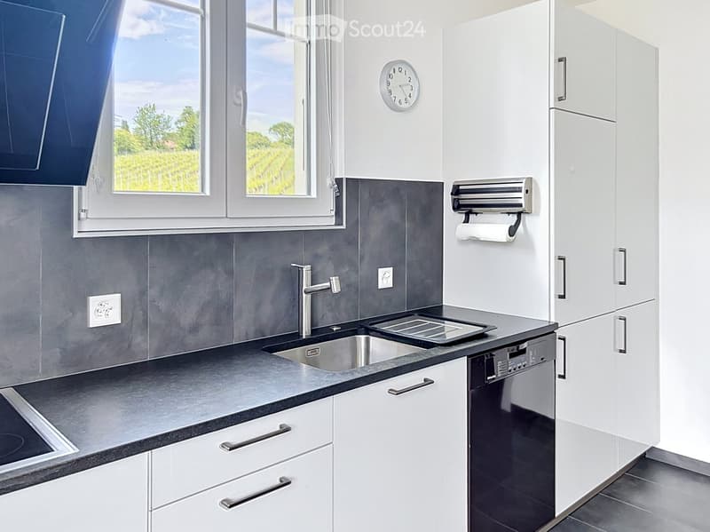 Modern kitchen with white cabinets, dark grey countertops, stainless steel sink and faucet, dishwasher, and a clock on the wall. Large windows offer a view of greenery outside.