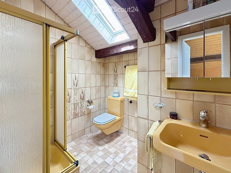 Bathroom with beige tiles, a toilet, a sink, a mirror, and a shower area with a glass door. A towel hangs on the side of the sink, and a small decorative item sits on the toilet tank.