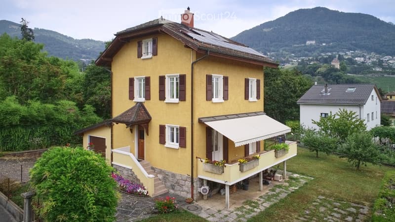 Two-story yellow house with white-framed windows, brown shutters, and a balcony with flower boxes. Solar panels on the roof and a scenic mountain view in the background.