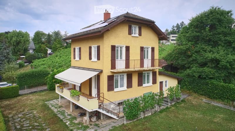 Two-story yellow house with brown shutters, multiple windows, a balcony, and a small garden area with plants and a staircase leading up to the house.