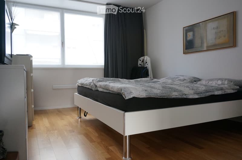 Modern bedroom with a white bed frame, grey bedding, wooden floor, large window with dark curtains, nightstand, and a framed picture on the wall.