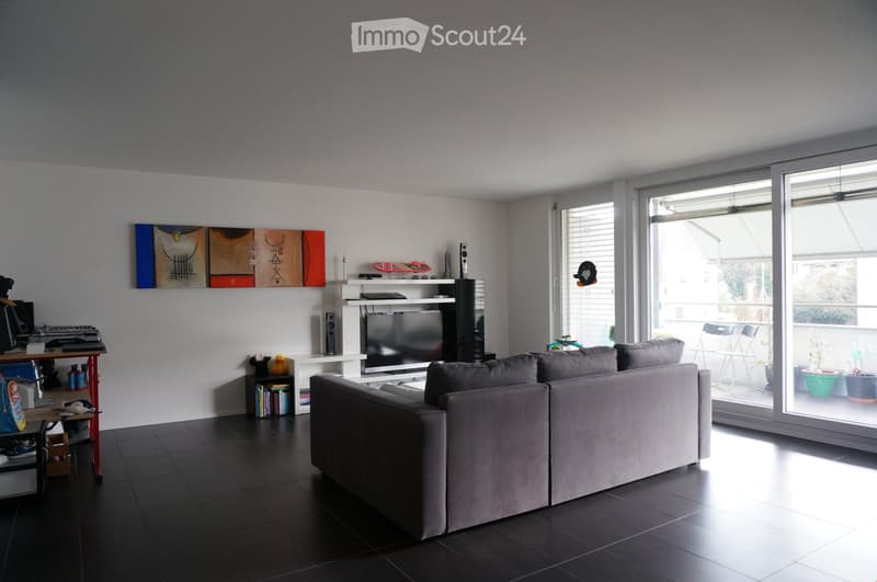 Modern living room with a grey sectional sofa, dark tiled floor, large windows, TV, shelves with books and decorative items, and a small table with a chair.
