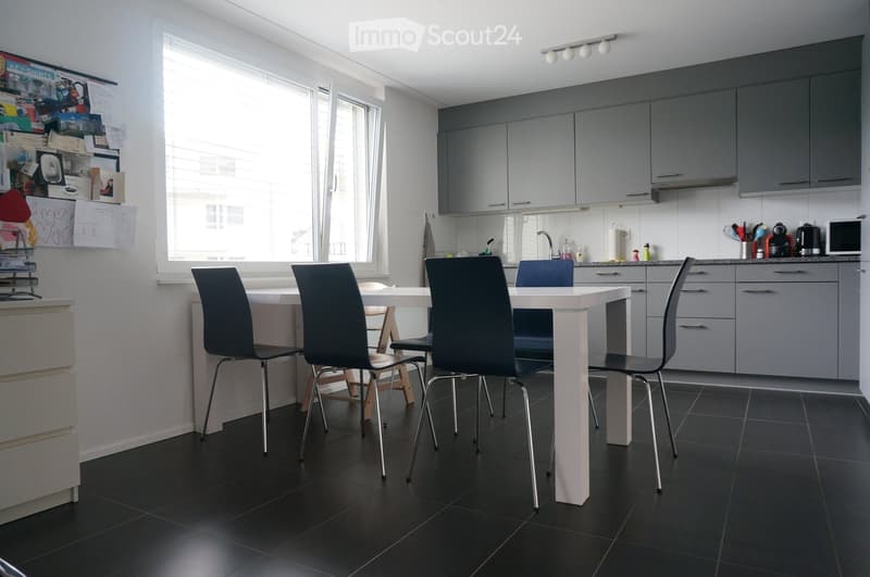 Modern kitchen with grey cabinets, white dining table, four black chairs, and various appliances including a microwave and a faucet.