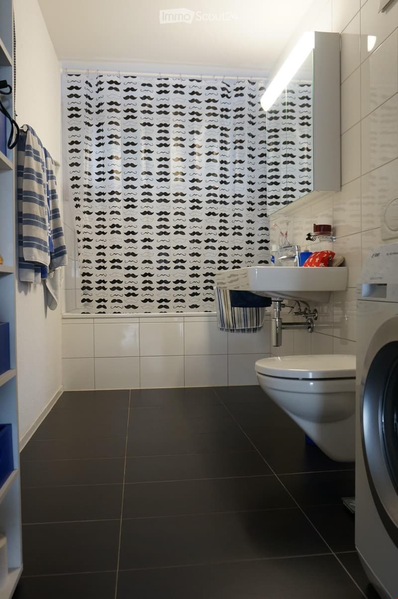 Modern bathroom with dark tiled floor, white tiled walls, bathtub, toilet, sink, mirror, and decorative shower curtain with mustache pattern.