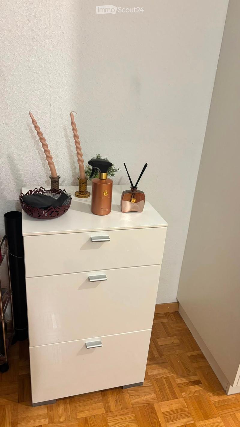 A small white cabinet with three drawers is placed on a wooden floor. On top of the cabinet, there are decorative items including candles, a spray bottle, incense sticks, and a decorative bowl.