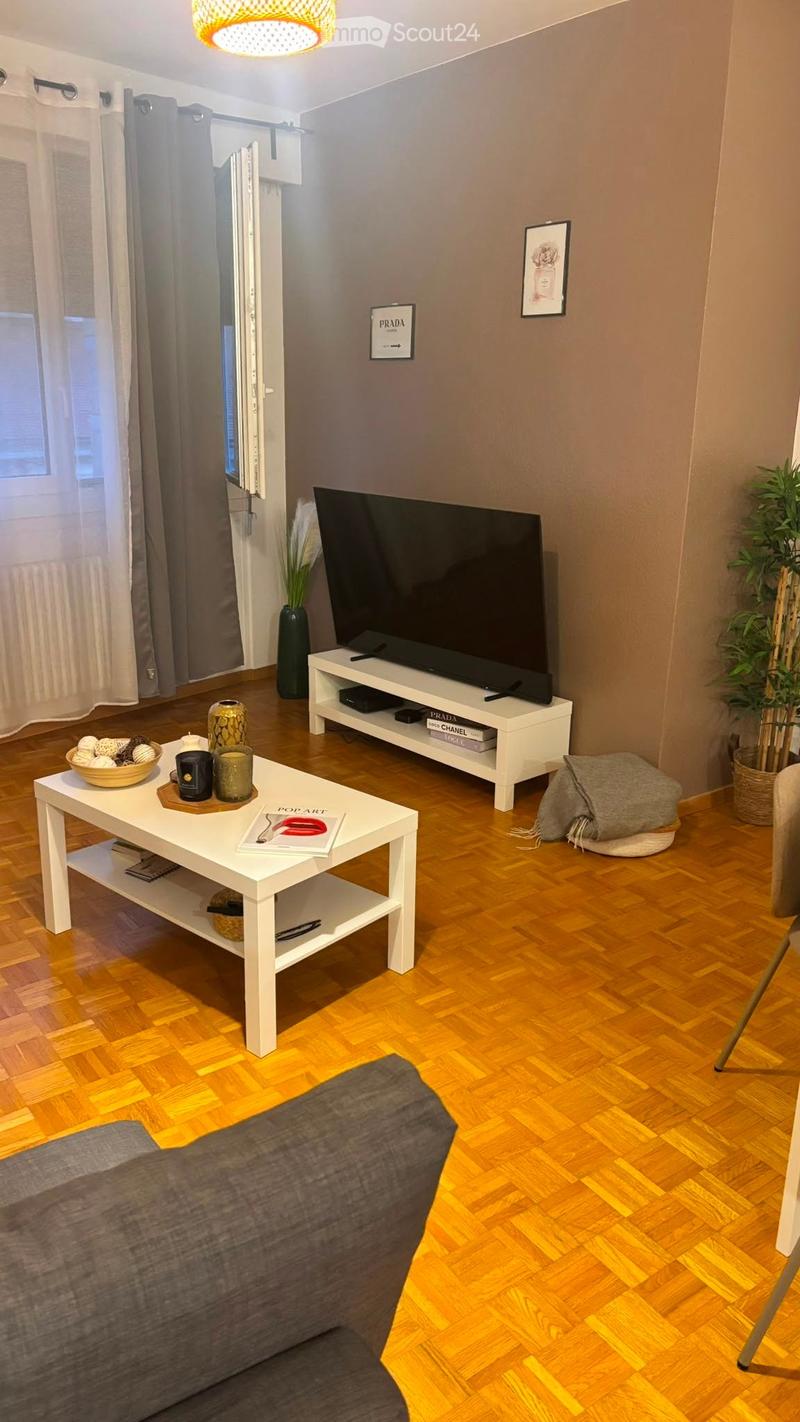 Modern living room with wooden flooring, a grey sofa, a white coffee table, a television on a white stand, and decorative items including a vase, plant, and picture frames.