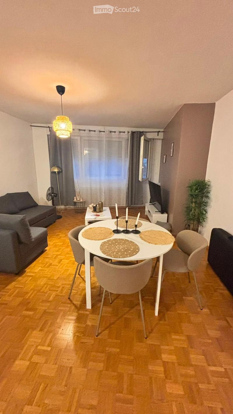 A modern living room with a grey sofa, a white dining table with four chairs, a television, a floor lamp, and a small fan. The room features wooden flooring and large windows with curtains.