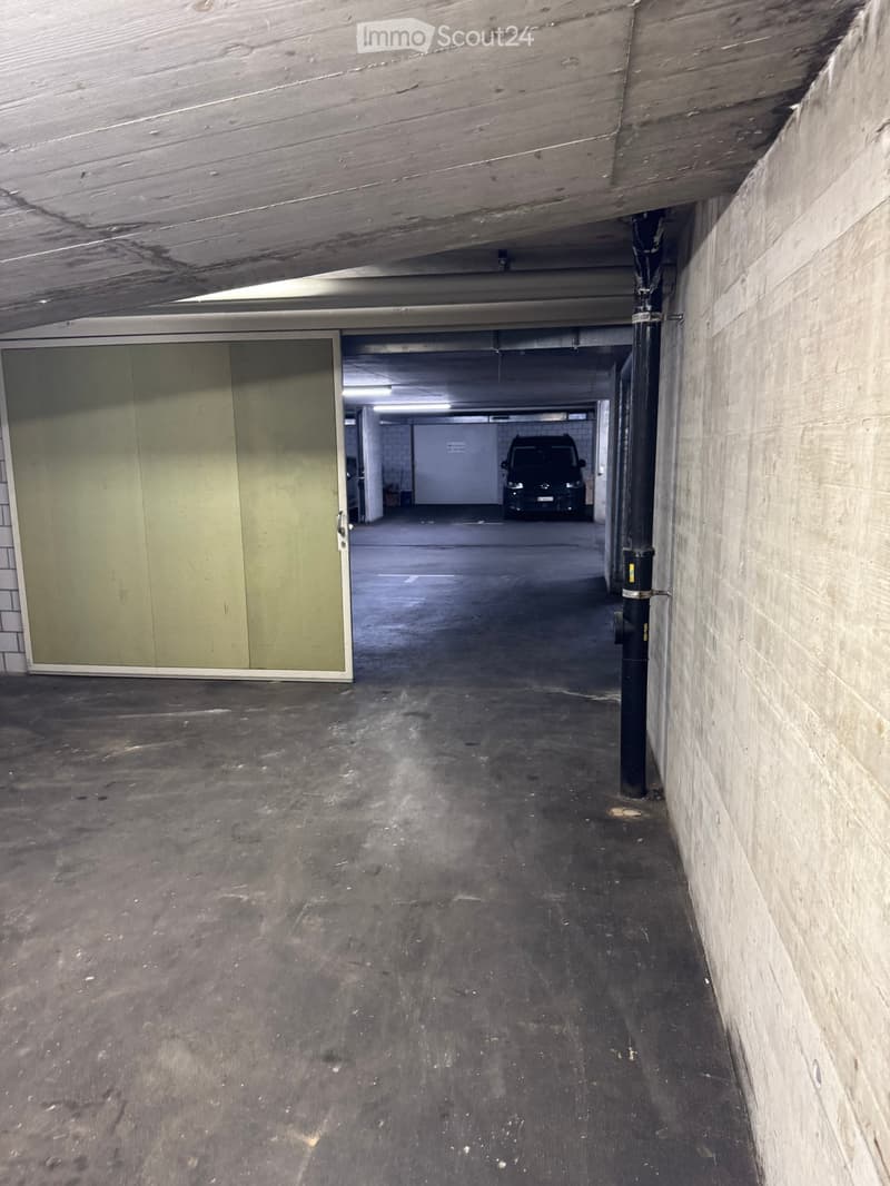 Empty underground parking corridor with concrete floors and walls, a closed garage door on the left, and a parked vehicle visible in the background.