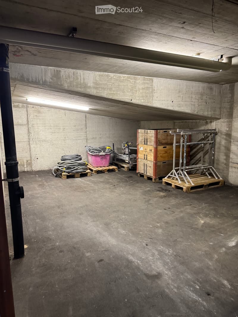 Empty underground space with concrete walls and floor, storage items including cables, boxes, and a metal rack on pallets.