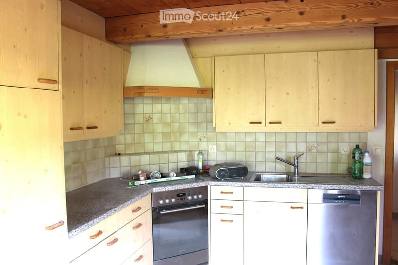 The kitchen features light wooden cabinets, a granite countertop, and stainless steel appliances including an oven, sink, and dishwasher. A wooden ceiling and tiled backsplash add warmth to the space.