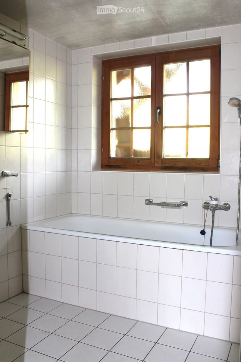 Bathroom with a white tiled bathtub, wooden-framed window, and modern fixtures including a handrail and showerhead.