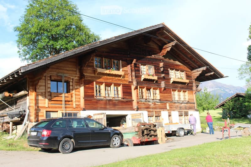 A large, traditional wooden house with multiple windows and balconies, surrounded by greenery and mountains. A car is parked in front, and there is a trailer and some construction materials nearby.