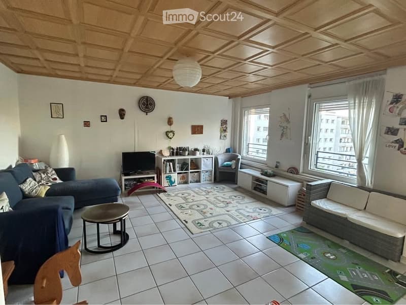 Living room with a blue sectional sofa, coffee table, armchair, shelf, TV, and decorative items. The room features a tiled floor and large windows with curtains.