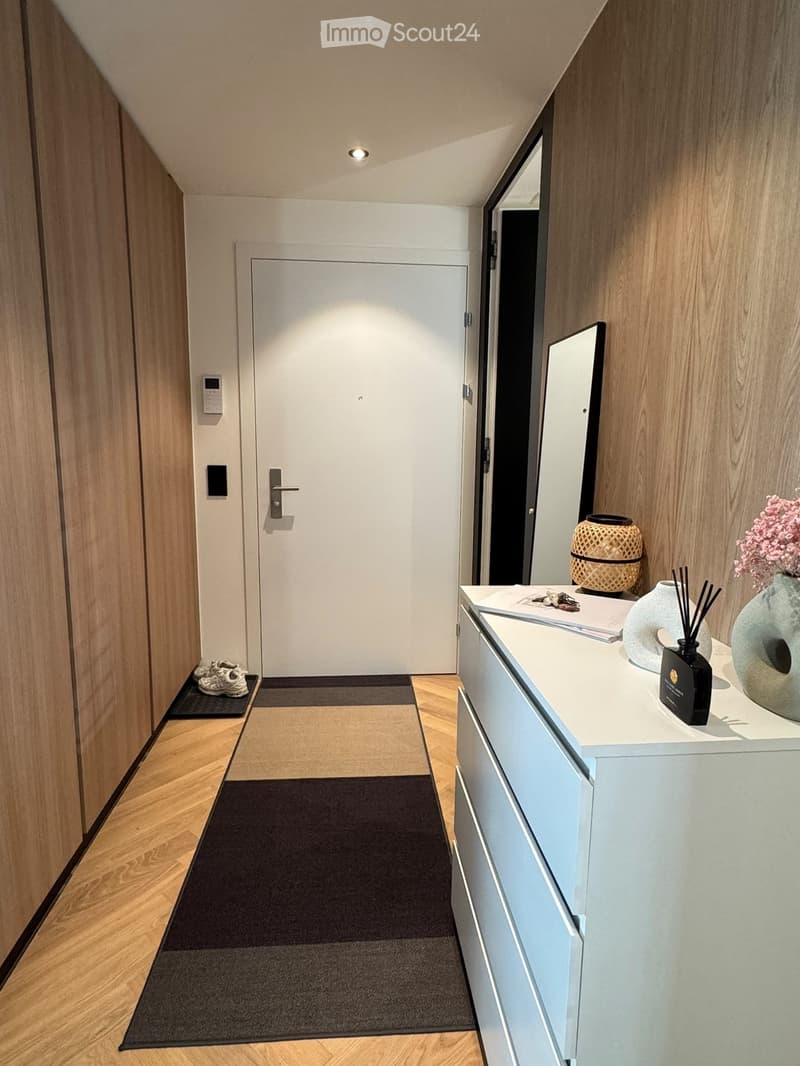 Modern hallway with wooden walls, herringbone flooring, a white door, a small console table with decorative items, and a pair of shoes on a mat.