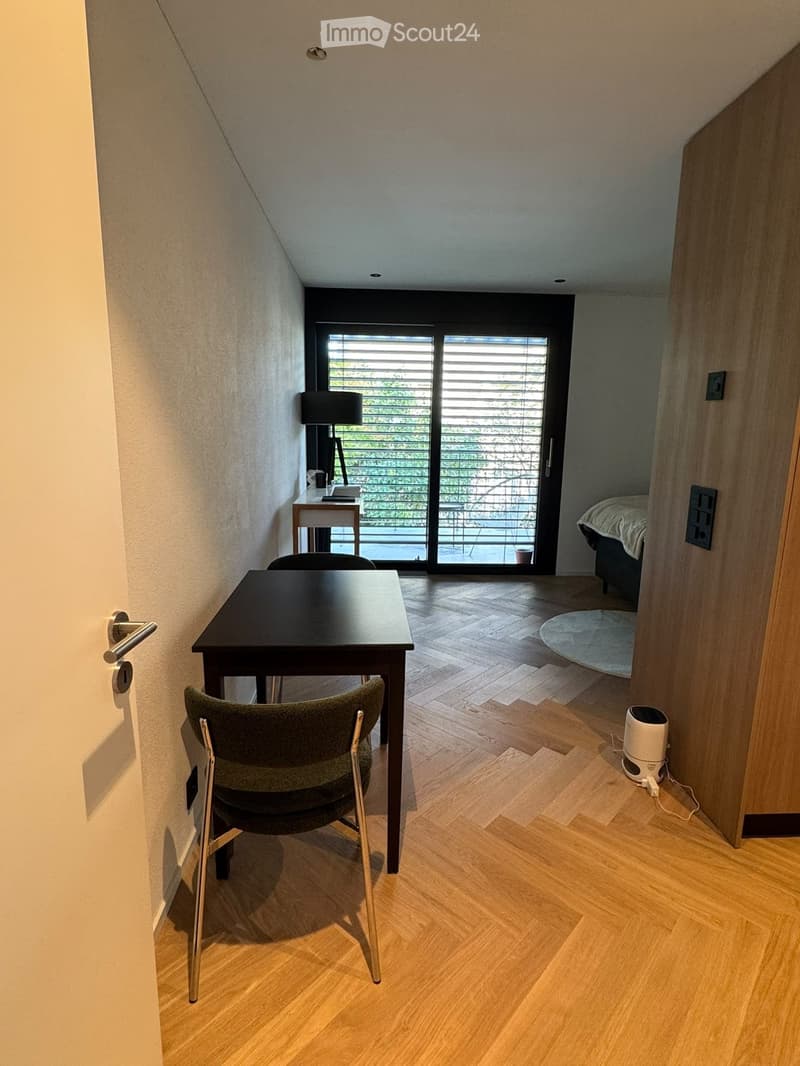 A modern, compact room with wooden flooring, a small desk with a lamp, a dining table with chairs, and a bed visible in the background. The room features large windows with blinds and a sliding glass door leading outside.