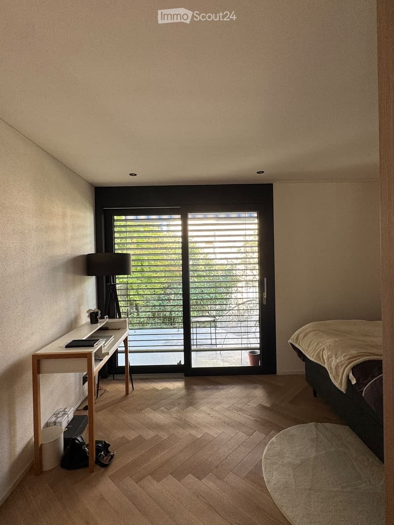 Modern bedroom with wooden flooring, large window with blinds, bed, desk, lamp, and small balcony area visible through the window.