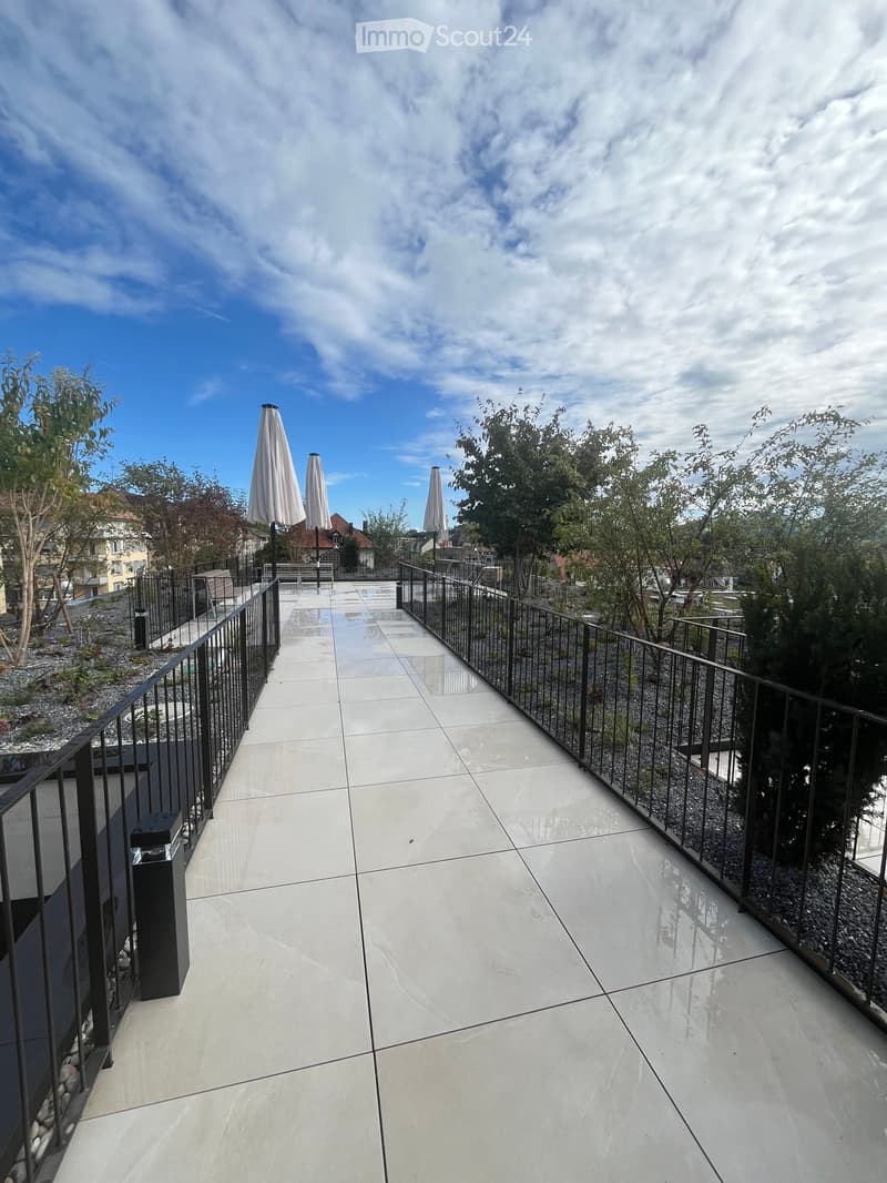 Modern balcony with white tiled flooring, black metal railing, and closed umbrellas in the background.