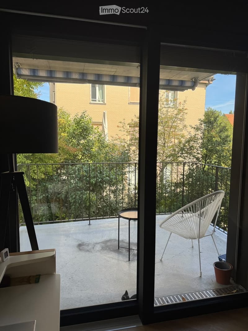 Balcony with a white modern chair, small round table, and a potted plant, overlooking a green area with trees and neighboring buildings.