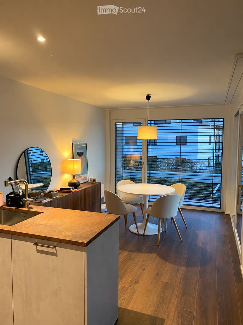 Modern kitchen with wooden countertops, white dining table, four chairs, and a sink area with a faucet. The space features a large window with blinds and a view of the outside.
