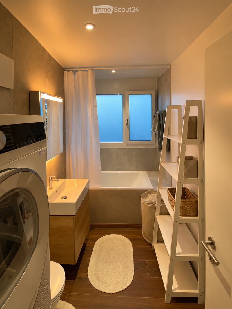 Modern bathroom with a bathtub, sink, toilet, washing machine, and a shelf with baskets. Features a large window with a curtain, wooden flooring, and a white rug.
