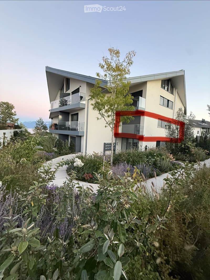 Modern two-story building with a sleek design, featuring large windows, balconies, and a landscaped garden area with various plants and shrubs.