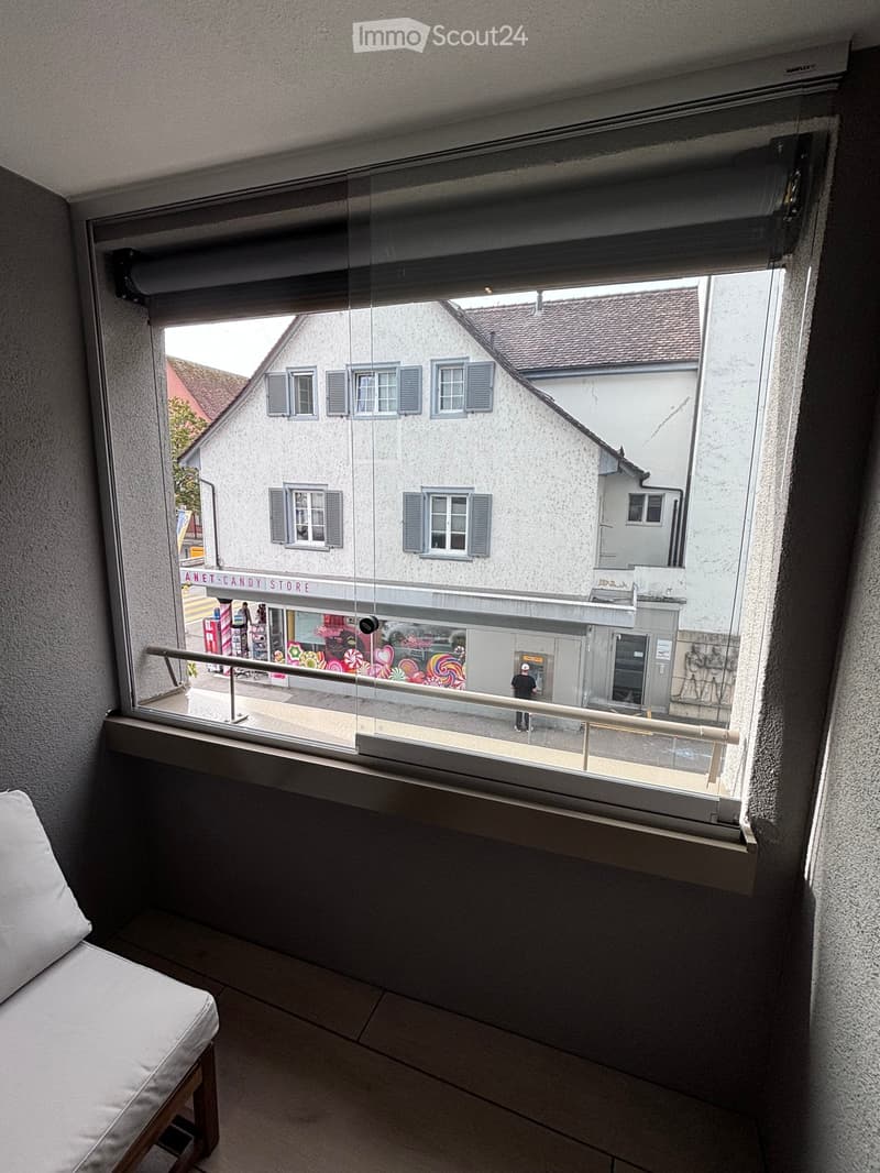 Balcony with a view of a street and buildings, featuring a white sofa and a glass railing.