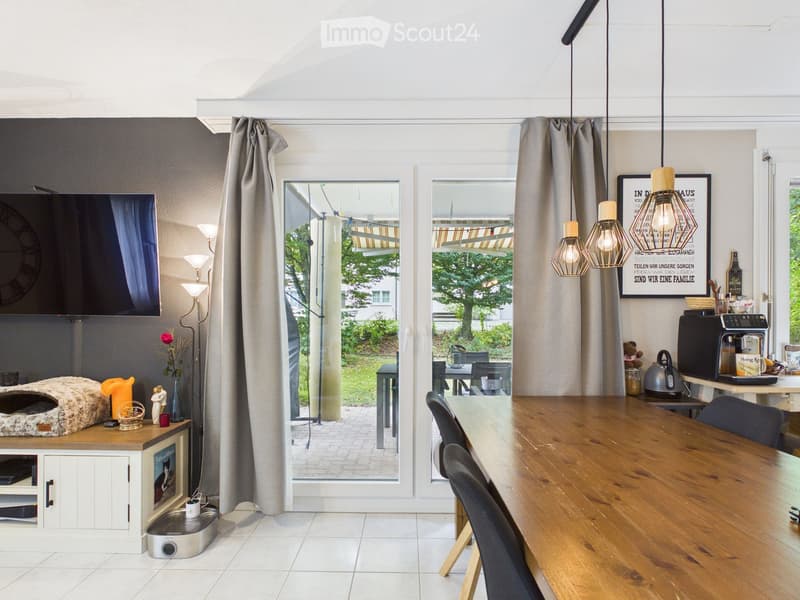 Bright living and dining area with large windows and sliding doors leading to a patio. Features a wooden dining table with chairs, a sofa, a television, a cat litter box, and a coffee machine.
