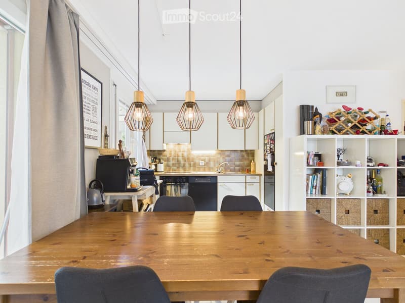 Open-plan kitchen and dining area with a wooden dining table, chairs, modern kitchen cabinets, appliances, and pendant lights.