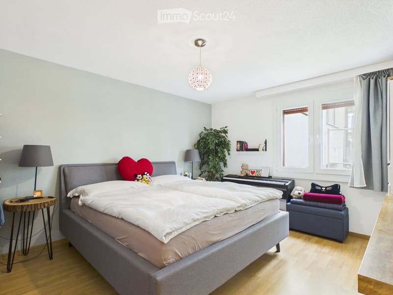 A modern bedroom with a large bed, a wooden stool, a small table with a lamp, a plant, and a shelf with books and a piano.