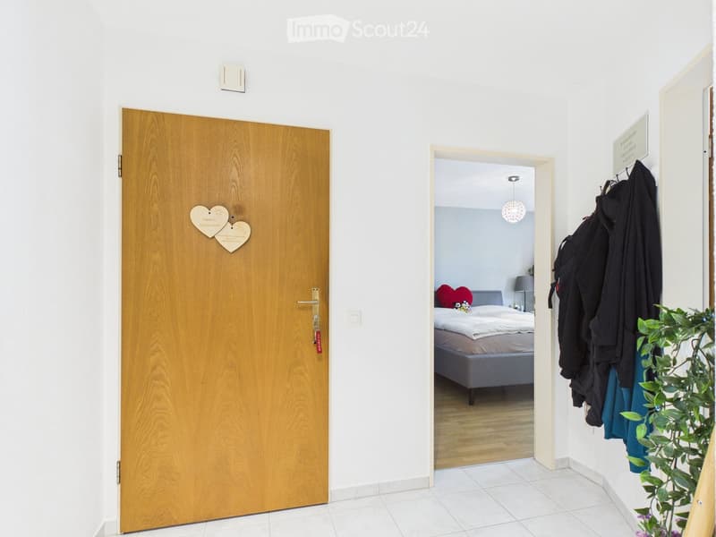 A hallway with a wooden door on the left and an open doorway leading to a bedroom on the right. The bedroom contains a bed with a red heart-shaped pillow, a nightstand, and a lamp. Clothes are hanging on a rack, and a potted plant is visible near the door