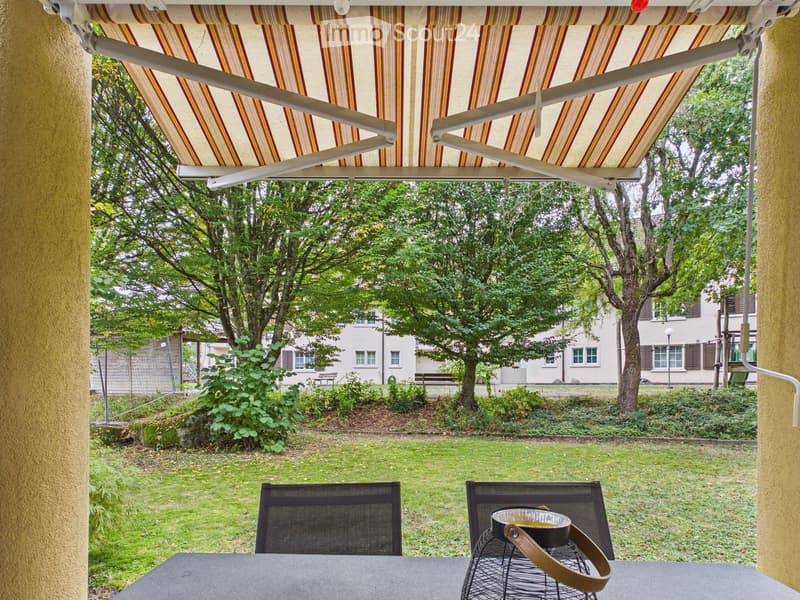 Balcony with a striped awning, a table, two chairs, and a small pot on a wire stand, overlooking a green garden area with trees and a building in the background.