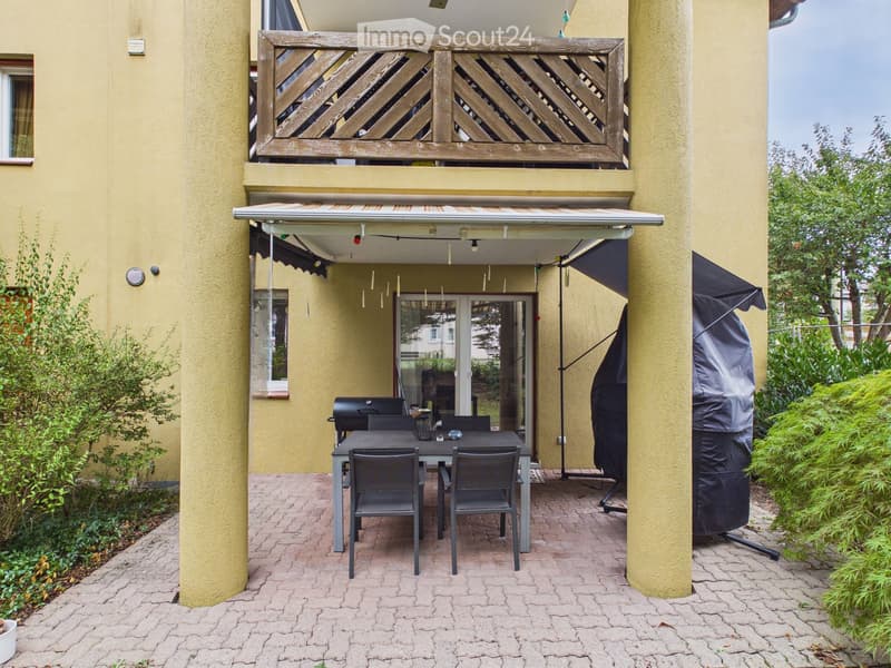 Outdoor patio with a table, chairs, and grill, covered by a retractable awning, surrounded by greenery and a wooden balcony above.
