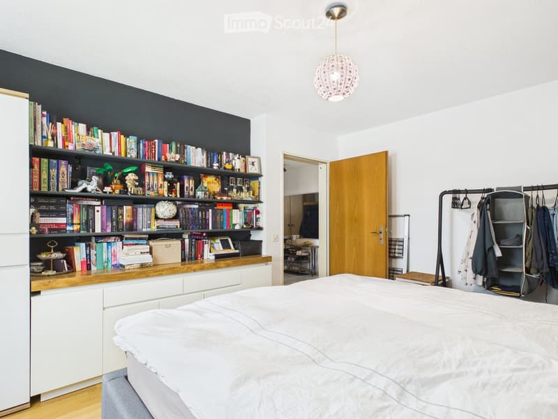 Modern bedroom with a large bed, built-in bookshelf filled with books and decorative items, a hanging pendant light, and a coat rack with jackets.