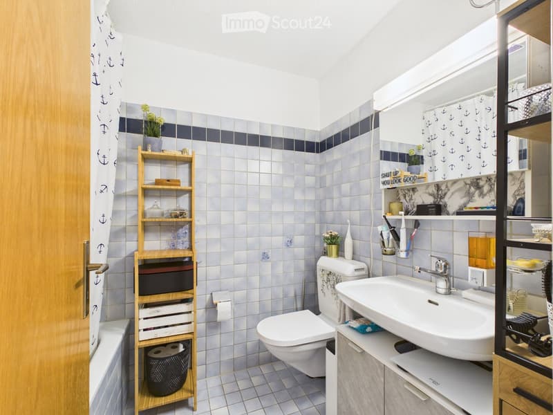 Modern bathroom with white tiles, a wooden shelf, a sink, a toilet, a bathtub, and a mirror. The decor includes small flower pots and a decorative sign.