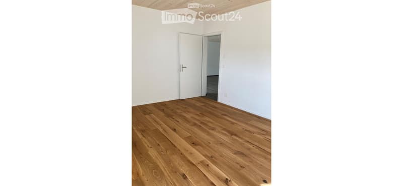 Empty room with wooden flooring and white walls, featuring a door leading to another space.