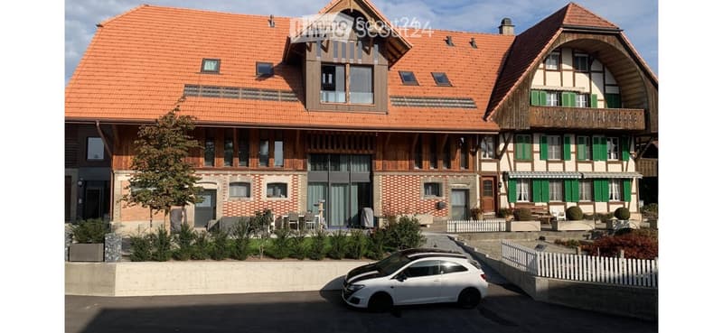 Two-story house with red-tiled roof, wooden facade, multiple windows, and a small garden area with outdoor seating.